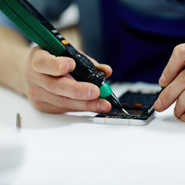 Closeup shot of male hands working on disassembling circuit board in mobile phone device and checking electric current with multimeter tool on table in maintenance shop with assorted tools